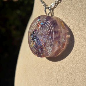Könnte beinhalten: Eine runde Anhängerhalskette mit einer silbernen Kette. Der Anhänger ist ein durchscheinendes Lila mit einem Sonnen- und Sterndesign. Die Sonne hat ein lächelndes Gesicht. Der Anhänger hat einen silbernen Rand mit Sternen und anderen himmlischen Designs.