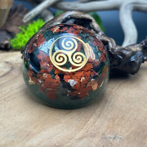 Peut inclure: Une sphère décorative avec une base vert foncé, parsemée d'accents rouges et dorés. Un symbole de triple spirale de couleur or est centré sur la sphère. La sphère est exposée sur une surface en bois.