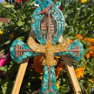 Könnte beinhalten: Ankh-Anhänger mit türkis-goldenem Design. Der Ankh hat ein türkisfarbenes Kreuz mit Kupferdrahtakzenten und einem goldfarbenen Vogel-Emblem. Der Hintergrund umfasst bunte Blumen und einen Holzständer.