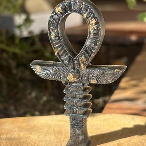 Può includere: Un ciondolo ankh grigio scuro con dettagli in foglia d'oro. L'ankh presenta un anello nella parte superiore, una barra orizzontale e una base verticale con un design a strati. Le ali sono spiegate sulla barra orizzontale.