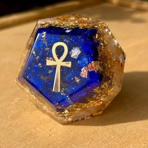 Peut inclure: Objet en résine géométrique à multiples facettes, avec un centre bleu profond et des accents dorés. Un symbole ankh doré est centré sur le bleu. Les bords extérieurs sont incrustés de feuilles d'or et d'écailles d'argent, créant un effet scintillant.