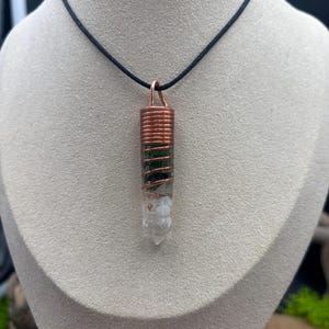 Pode incluir: Um colar com um pingente de cristal envolto em fio de cobre, com um cristal verde e branco. O colar está em um cordão preto.