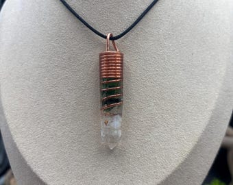 Puede incluir: Un collar con un colgante de cristal envuelto en alambre de cobre, con un cristal verde y blanco. El collar está en un cordón negro.