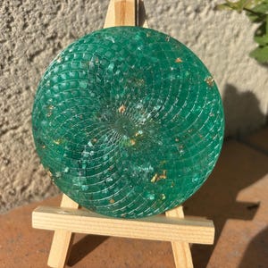Könnte beinhalten: Ein runder, smaragdgrüner Dekorationsgegenstand mit einem Spiralmuster, präsentiert auf einer kleinen Holzstaffelei. Das Stück hat eine strukturierte Oberfläche mit Goldflocken, die einen einzigartigen Effekt erzeugt.