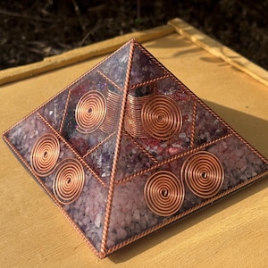 Op de afbeelding: Een piramidevormig orgonite-apparaat met een koperdraadframe en spiraalaccenten. De binnenkant van de piramide bevat een mix van paarse en rode stenen, ingekapseld in heldere hars. De koperen spiralen zijn gelijkmatig verdeeld.