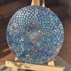 Peut inclure: Une œuvre d'art circulaire en mosaïque avec un motif en spirale. L'œuvre combine des carreaux bleus et turquoise, avec des accents dorés. Elle est présentée sur un petit chevalet en bois, mettant en valeur le design complexe et les variations de couleurs.