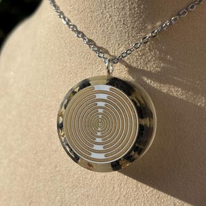 Peut inclure: Un collier à chaîne argentée avec un pendentif circulaire. Le pendentif présente un motif en spirale concentrique or et blanc, enfermé dans de la résine transparente. La résine est entourée d'un motif tacheté noir et or.