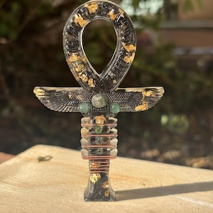 Può includere: Ciondolo a forma di Ankh in resina con inserti in foglia d'oro. L'Ankh, simbolo di vita, presenta un'ansa, una barra trasversale e un gambo verticale. Le ali sono dettagliate in nero e oro. Un gioiello unico.