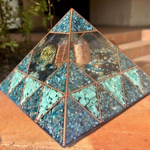 Puede incluir: Una pirámide decorativa con marco de cobre y detalles turquesa y azul. El interior de la pirámide presenta una espiral dorada y estructuras de cobre. La pirámide se exhibe sobre una superficie de ladrillo.