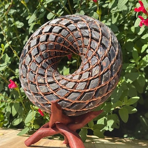 Könnte beinhalten: Eine einzigartige, donut-förmige Skulptur mit einer dunklen, strukturierten Oberfläche, die kunstvoll mit Kupferdraht verwoben ist. Die Skulptur steht auf einem rotbraunen Holzständer vor grünem Laub und roten Blüten.