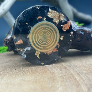 Könnte beinhalten: Ein schwarzer Harz-Untersetzer mit goldenen und kupfernen Flocken und einem grünen und goldenen geometrischen Design in der Mitte.
