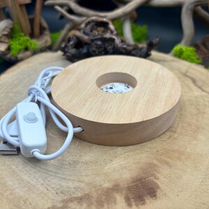 May include: A wooden round base with a circular cutout in the center. The base has a white cord with a switch and a USB plug. The base is on a wooden surface.