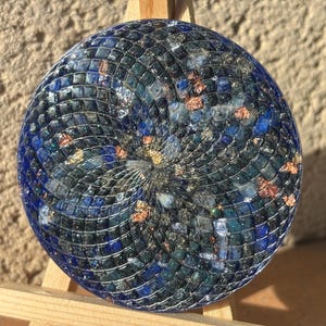 Könnte beinhalten: Ein rundes dekoratives Mosaikstück mit einem Spiralmuster. Es zeigt dunkelblaue, teal- und kupferfarbene Elemente, die ein strukturiertes Aussehen erzeugen. Das Mosaik wird auf einer kleinen Holzstaffelei präsentiert. Der Durchmesser beträgt ca. 15 cm.