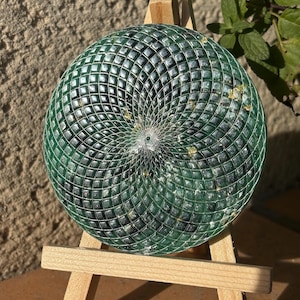 Könnte beinhalten: Ein grünes, kreisförmiges Dekorationsobjekt mit einem geometrischen Muster, das auf einer kleinen Holzstaffelei präsentiert wird. Das Objekt hat ein Spiraldesign mit gitterartiger Textur und einem zentralen Punkt. Goldflocken sind sichtbar.