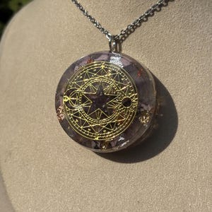 Peut inclure: Un collier pendentif rond avec une chaîne argentée. Le pendentif présente un motif géométrique doré sur une résine violette et dorée. Le collier est présenté sur une surface aux tons neutres.