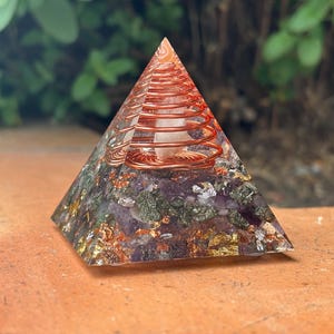 Peut inclure: Une pyramide énergétique orgone. La base est un mélange de matériaux violets, verts et dorés. Une spirale de cuivre entoure un cristal clair au centre, avec un sommet teinté d'orange.