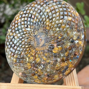 Könnte beinhalten: Ein dekoratives, rundes Kunstwerk aus Harz mit einem Spiralmuster. Das Kunstwerk zeigt ein Mosaikmuster aus kleinen, quadratischen Fliesen in Gold-, Braun- und Klartönen mit eingebetteten Metallflocken. Das Stück wird auf einer kleinen Holzstaffelei ausgestellt.