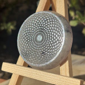 Puede incluir: Un objeto redondo plateado con un patrón geométrico detallado. El diseño presenta círculos concéntricos de triángulos que irradian desde el centro. El objeto se muestra sobre un pequeño caballete de madera, con un fondo verde borroso.