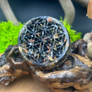 May include: A black resin coaster featuring a gold and silver flower of life design. The coaster is round and has a raised edge.