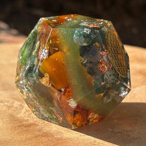 May include: A faceted, geometric resin sculpture with a mix of green, orange, and gold leaf inclusions. The sculpture has a golden symbol on one side. The object is sitting on a wooden surface.