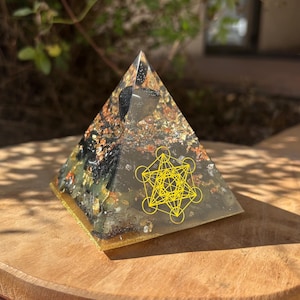 Op de afbeelding: Een helder, piramidevormig kunstwerk van hars met ingebedde stenen en goudvlokken. De piramide heeft een geel Metatron's Cube-symbool aan de voorkant. De basis is goudkleurig en de piramide staat op een houten oppervlak.