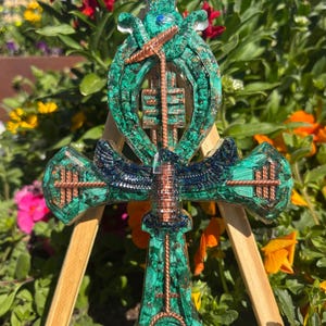 Peut inclure: Une sculpture détaillée d'une croix Ankh verte et cuivrée. La croix présente des motifs complexes et est exposée sur un petit chevalet en bois, sur fond de fleurs colorées. L'Ankh est un symbole de vie.