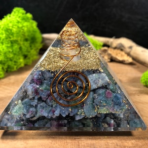 Könnte beinhalten: Eine klare Glaspyramide mit einer Kupferspirale im Inneren. Die Pyramide ist gefüllt mit kleinen blauen, grünen und violetten Kristallen und goldenem Glitzer.