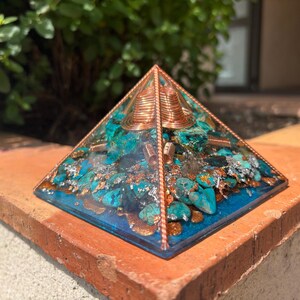 May include: A handcrafted pyramid with a copper frame, filled with turquoise stones, silver flakes, and resin. The pyramid has a copper coil at the top. The base is a deep blue color. The pyramid is displayed on a brick ledge.