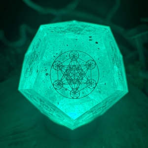 Puede incluir: Una lámpara geométrica brillante de color verde azulado con un diseño del Cubo de Metatrón grabado en su superficie. La lámpara está iluminada, proyectando un brillo suave y etéreo. El diseño es un patrón geométrico complejo.