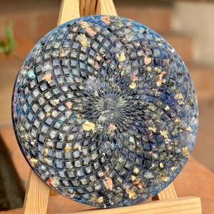 Könnte beinhalten: Eine runde, dekorative Resin-Platte in Blau- und Goldtönen. Sie weist ein geometrisches Muster und Goldblatt-Akzente auf. Präsentiert wird sie auf einem kleinen Holzstaffelei.