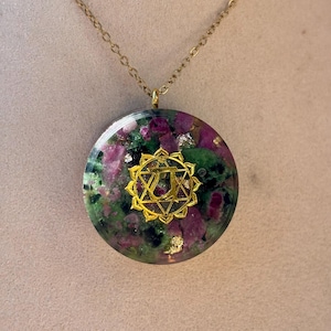 Peut inclure: Un collier pendentif circulaire avec une chaîne dorée et un disque en résine contenant des fragments de pierre verts et magenta incrustés avec des accents de feuilles d'or. Un symbole en métal doré est centré sur le devant du disque en résine.
