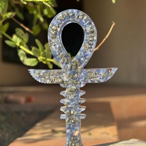 Peut inclure: Un pendentif ankh bleu et or, symbole de vie, est présenté. L'ankh comporte une boucle en haut, une barre horizontale et une tige verticale. Le pendentif est fait d'un matériau bleu translucide avec des paillettes dorées, sur un fond flou.