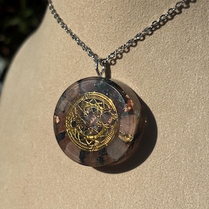 May include: A round pendant necklace with a silver chain. The pendant features a gold-colored, ornate design set within a clear resin, with brown and black flecks. The necklace is displayed on a neutral-toned surface.