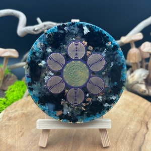 Pode incluir: Uma peça de arte de resina redonda, azul, preta e dourada com um design geométrico roxo e dourado. A peça está em um pequeno cavalete de madeira.
