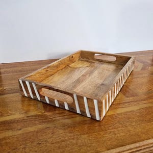 Peut inclure: Un plateau de service rectangulaire en bois avec des rayures blanches peintes et deux poignées. Le plateau est en bois naturel et a un aspect rustique.