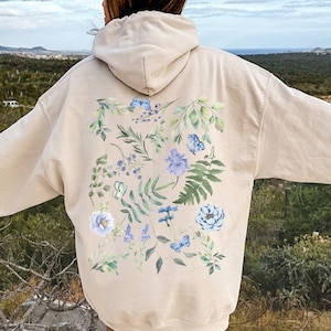 May include: A beige hoodie with a floral design on the back. The design features blue and green watercolor flowers and leaves.