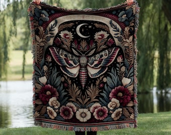 May include: A tapestry throw blanket with a dark background featuring a large moth with a crescent moon and stars above it. The moth is surrounded by floral designs in shades of red, pink, and white.