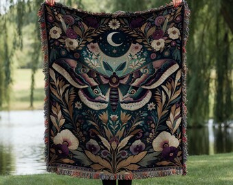 May include: A tapestry throw blanket with a dark green background featuring a large, colorful moth with intricate details. The moth is surrounded by floral patterns in shades of purple, pink, and white. A crescent moon with stars is visible in the upper center of the design.