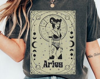 May include: A dark gray t-shirt with a graphic print of a woman with ram horns standing in front of a zodiac wheel. The graphic is in black and white with a beige background. The text "Aries" is printed below the graphic.