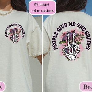 May include: White t-shirt with a graphic of a skeleton hand making a peace sign with mushrooms and flowers. The text "People Give Me The Creeps" is written around the hand.
