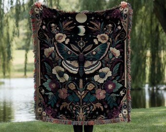 May include: A black throw blanket with a large moth and floral design. The moth is teal and white, surrounded by cream, teal, and purple flowers. The blanket has a fringed edge and is held up in front of a green background.