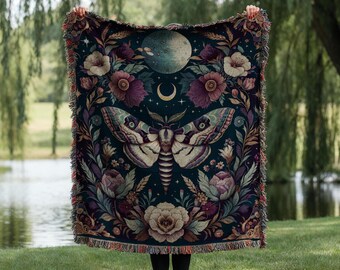 May include: A dark teal throw blanket with a moth and floral design. The blanket features a large moth in the center, surrounded by flowers, a crescent moon, and a planet. The edges have a fringed trim.