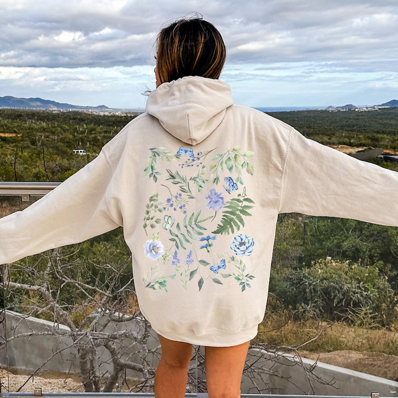 May include: A beige hoodie with a floral design on the back. The design features blue and green watercolor flowers and leaves.