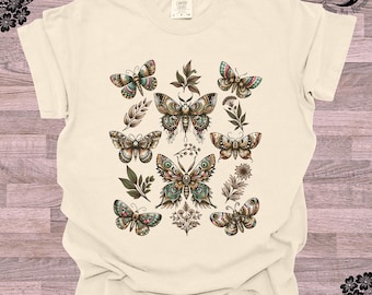 May include: A cream-colored t-shirt with a pattern of colorful butterflies and leaves. The butterflies are detailed with intricate patterns and have a vintage aesthetic.
