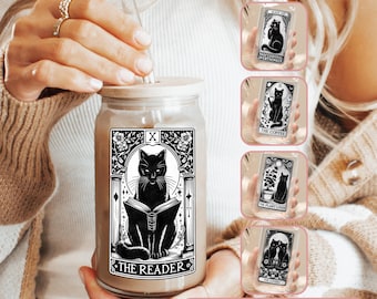 May include: A clear glass jar with a black cat reading a book design. The jar has a black and white design with the words "The Reader" and "X" on it. The jar has a lid and a straw. There are five different designs to choose from, including a black cat with the words "Professional Overthinker", a black cat with the words "The Coffee", a black cat with a plant and the words "The Plant Lover", and a black cat with the words "The Lovers".
