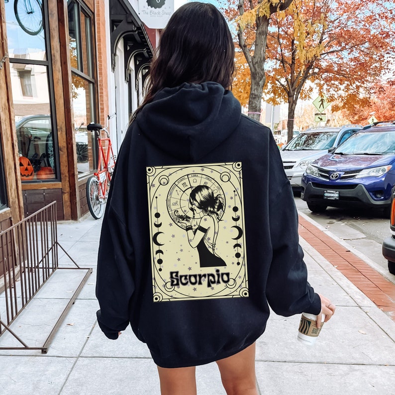 May include: Black hoodie with a graphic print of a woman with a scorpion and the word "Scorpio" in a yellow circle. The graphic is surrounded by a black frame.