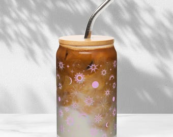 May include: A glass can with a bamboo lid filled with iced coffee. The can has a pink and gold celestial pattern with stars, moons, and suns.