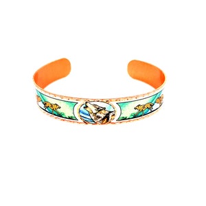 May include: Copper bracelet with a white enamel design featuring a howling wolf and two running wolves. The bracelet is a light orange color.