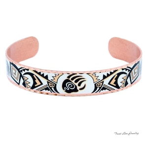 May include: Copper cuff bracelet with a Native American design. The bracelet features a white band with black, tan, and geometric patterns. A bear paw symbol is centered on the bracelet. The inside of the bracelet has a copper finish.