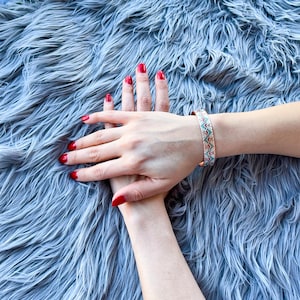 May include: Two hands with red nail polish on a gray fuzzy surface. One hand is wearing a colorful patterned bracelet.